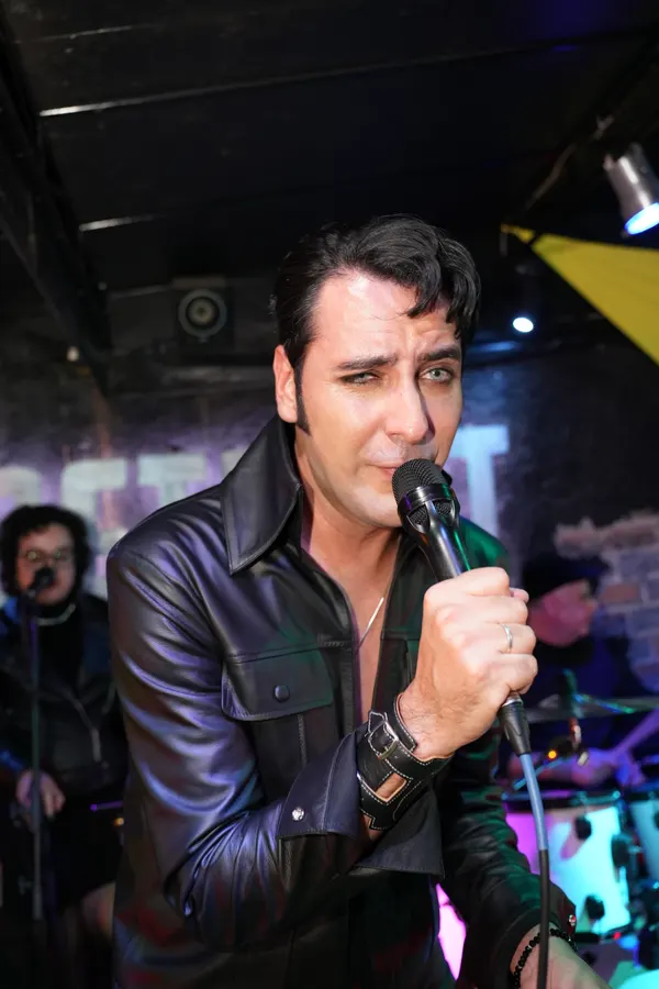 Caio Presley de jaqueta de couro preto, bem perto do microfone, com a baixista ao fundo