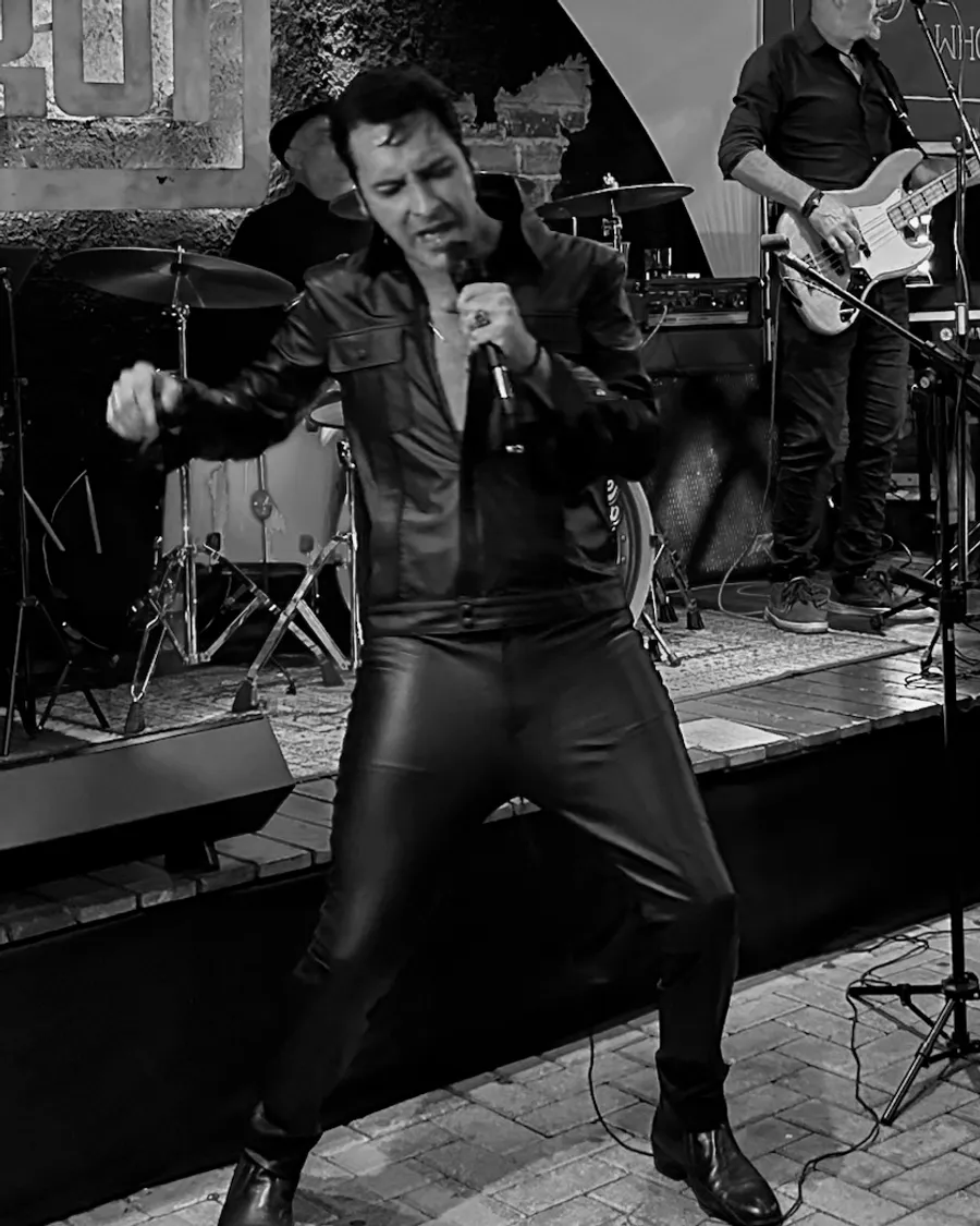 Caio Presley dançando no palco, de jaqueta e calça de couro preto, em foto preto e branco, com a bateria e o baixista ao fundo
